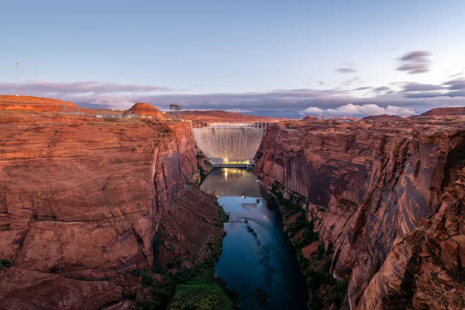 View of Glen Canyon Dam with vertical red stone cliffs on either side, Colorado River at bottom, and dusk sky in background.